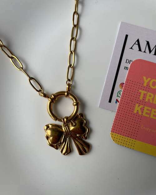 Gold necklace with a bow pendant on a white surface with a magazine in the background.