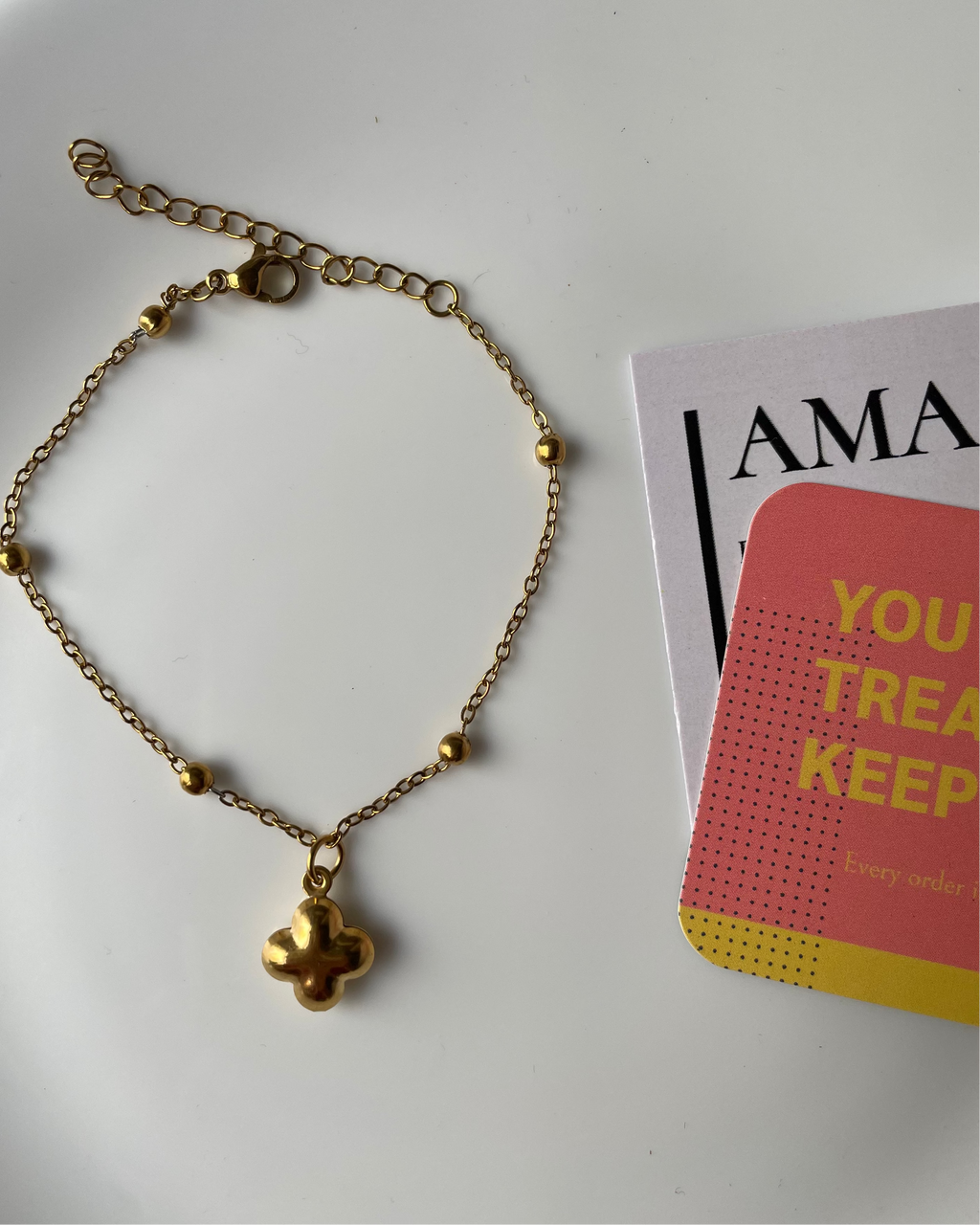 Gold bracelet with a clover-shaped pendant on a white surface next to a magazine.