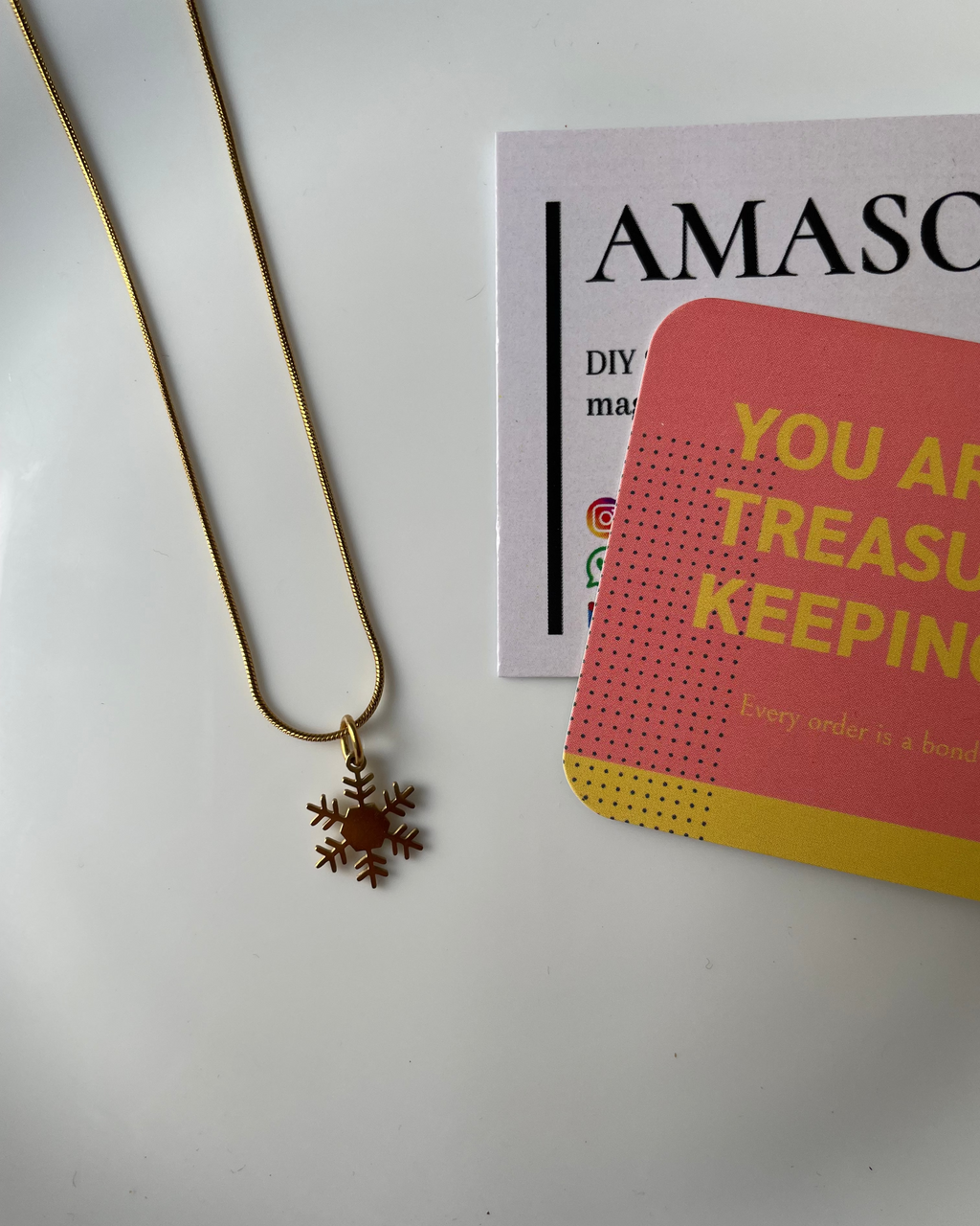 Gold necklace with a snowflake pendant on a white surface with Amazon promotional material.