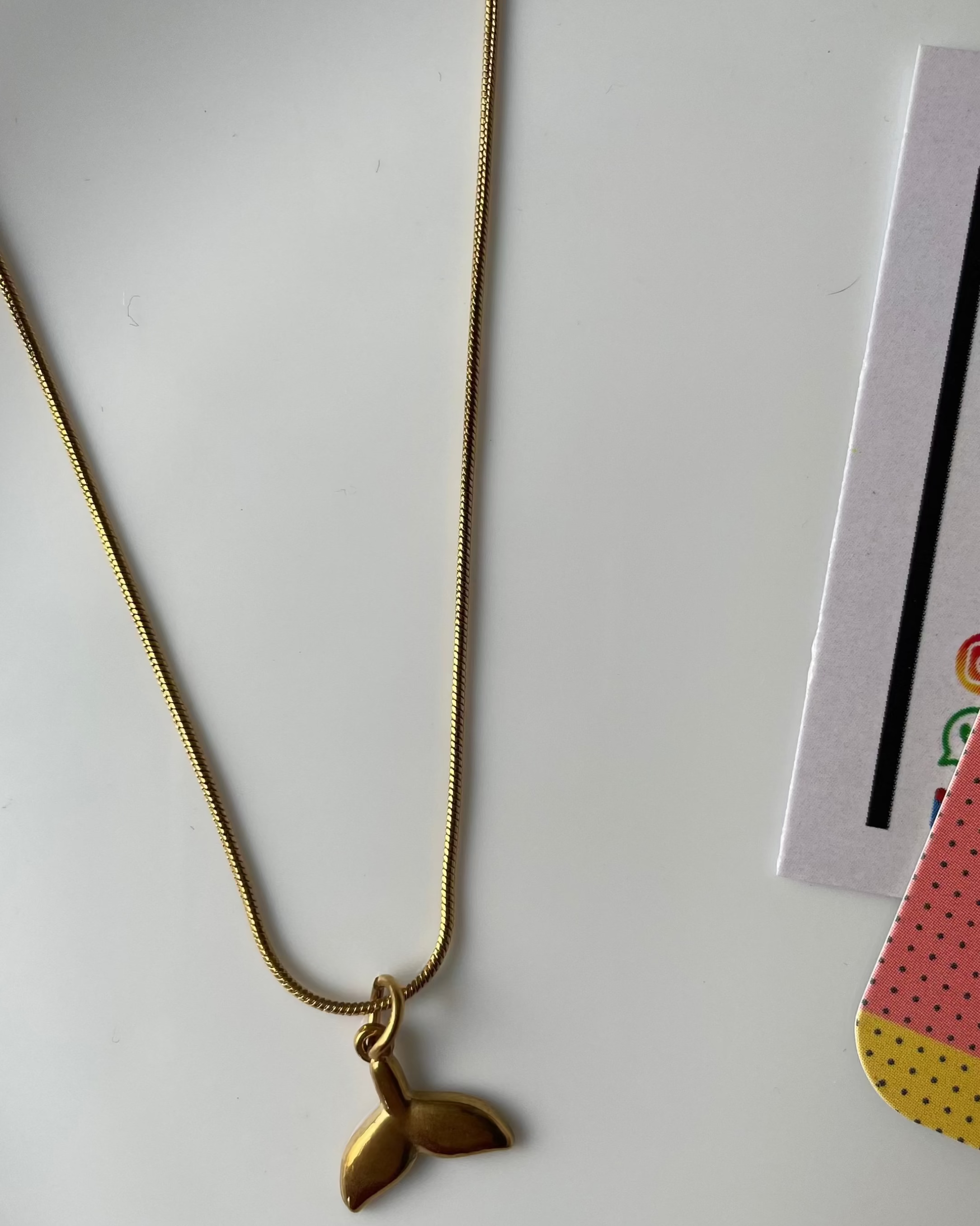 Gold necklace with a leaf pendant on a white surface with a magazine in the background.