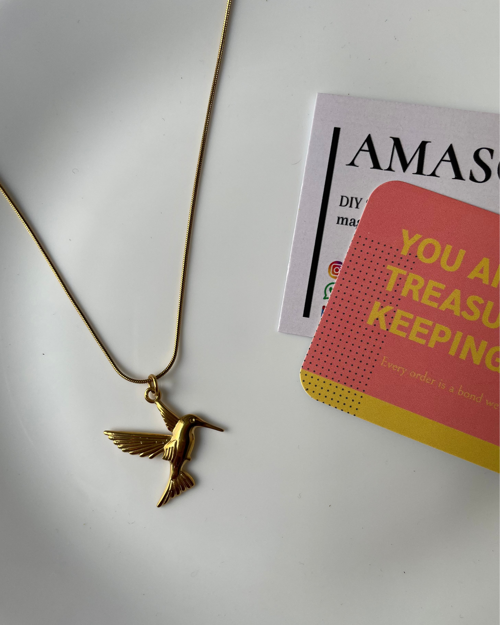 Gold hummingbird necklace on a white surface with a magazine in the background