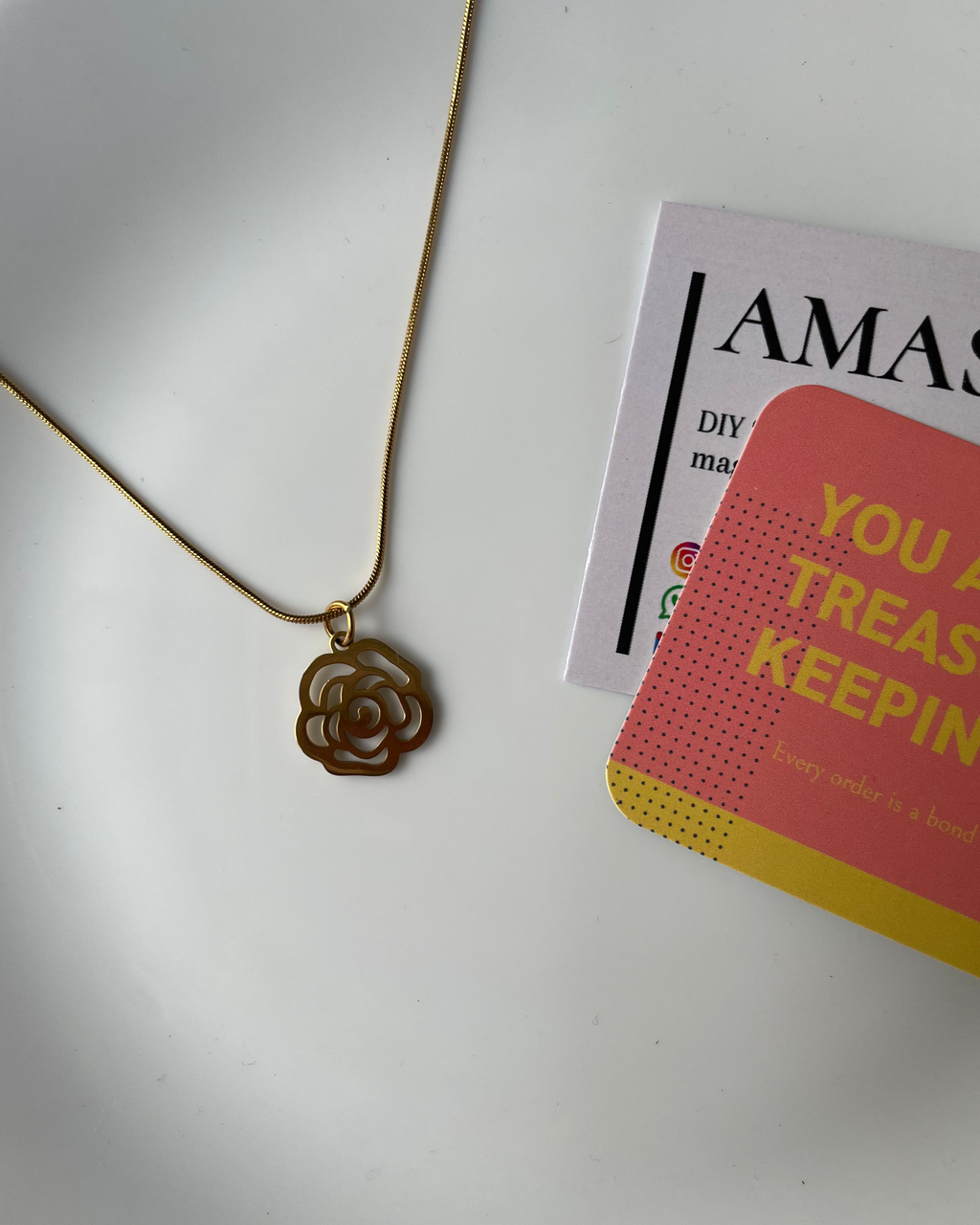 Gold necklace with a flower pendant on a white surface next to an Amazon magazine.