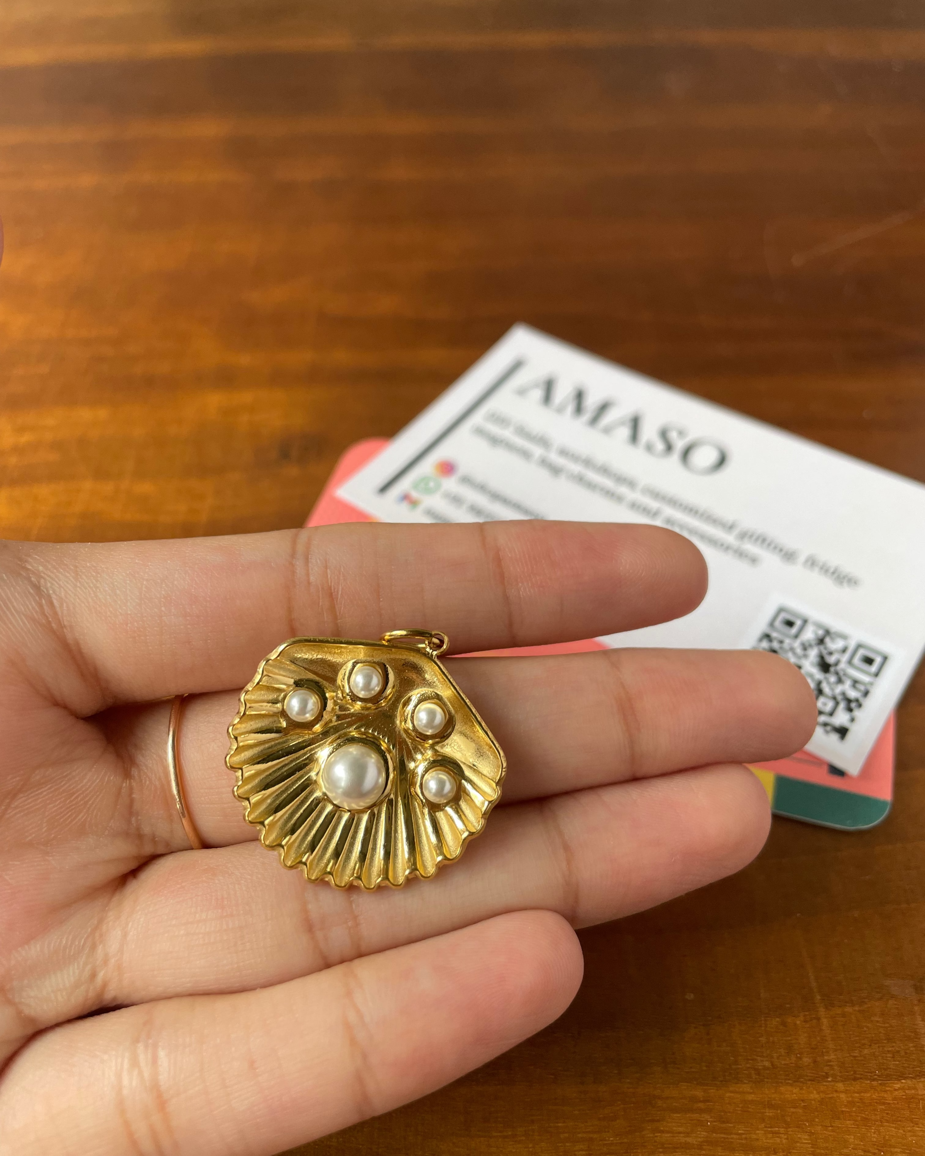 A gold shell-shaped pendant with pearl accents is held in a person's hand over a wooden table, with a branded card in the background.