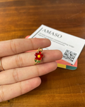 A hand holding a small gold ring shaped like a red and yellow flower with green leaves, resting on a wooden table next to an Amaso gift bag.