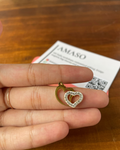 A gold heart-shaped pendant with a clear crystal border is held on a person's fingers over a wooden table, with a card in the background.