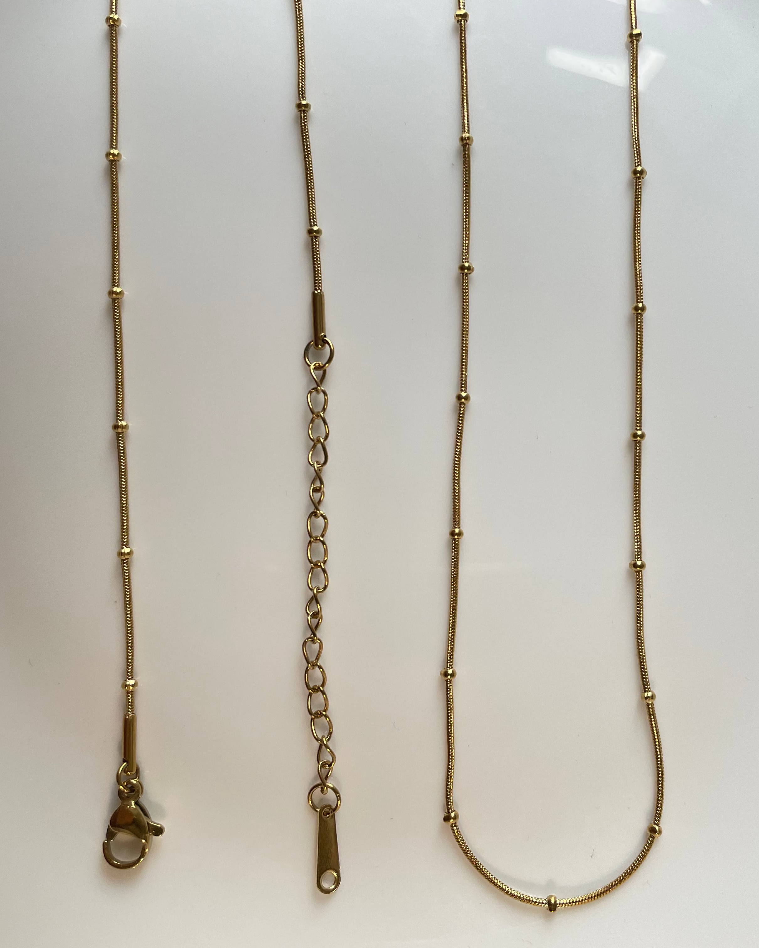 A gold-toned beaded chain necklace laid flat on a white surface, showing the full length of the chain and clasp from multiple angles.