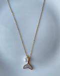 A delicate gold chain necklace featuring a mermaid tail pendant with a white mother-of-pearl inlay and a small white pearl bead, displayed on a light blue-gray surface.