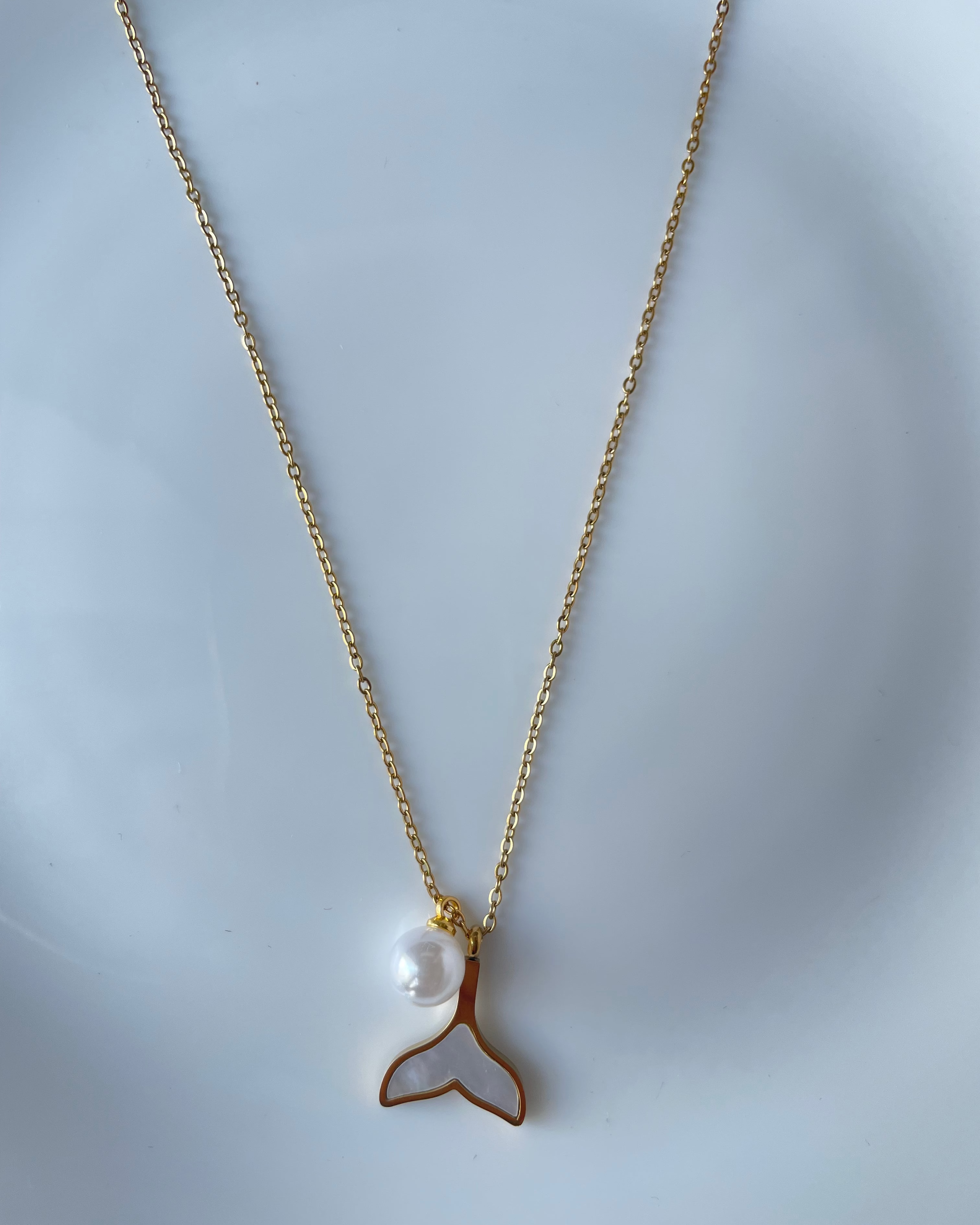 A delicate gold chain necklace featuring a mermaid tail pendant with a white mother-of-pearl inlay and a small white pearl bead, displayed on a light blue-gray surface.
