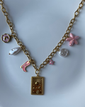 A gold-tone chain necklace with a rectangular pendant and several dangling charms, including pink and white pearl-like beads, displayed on a white plate.