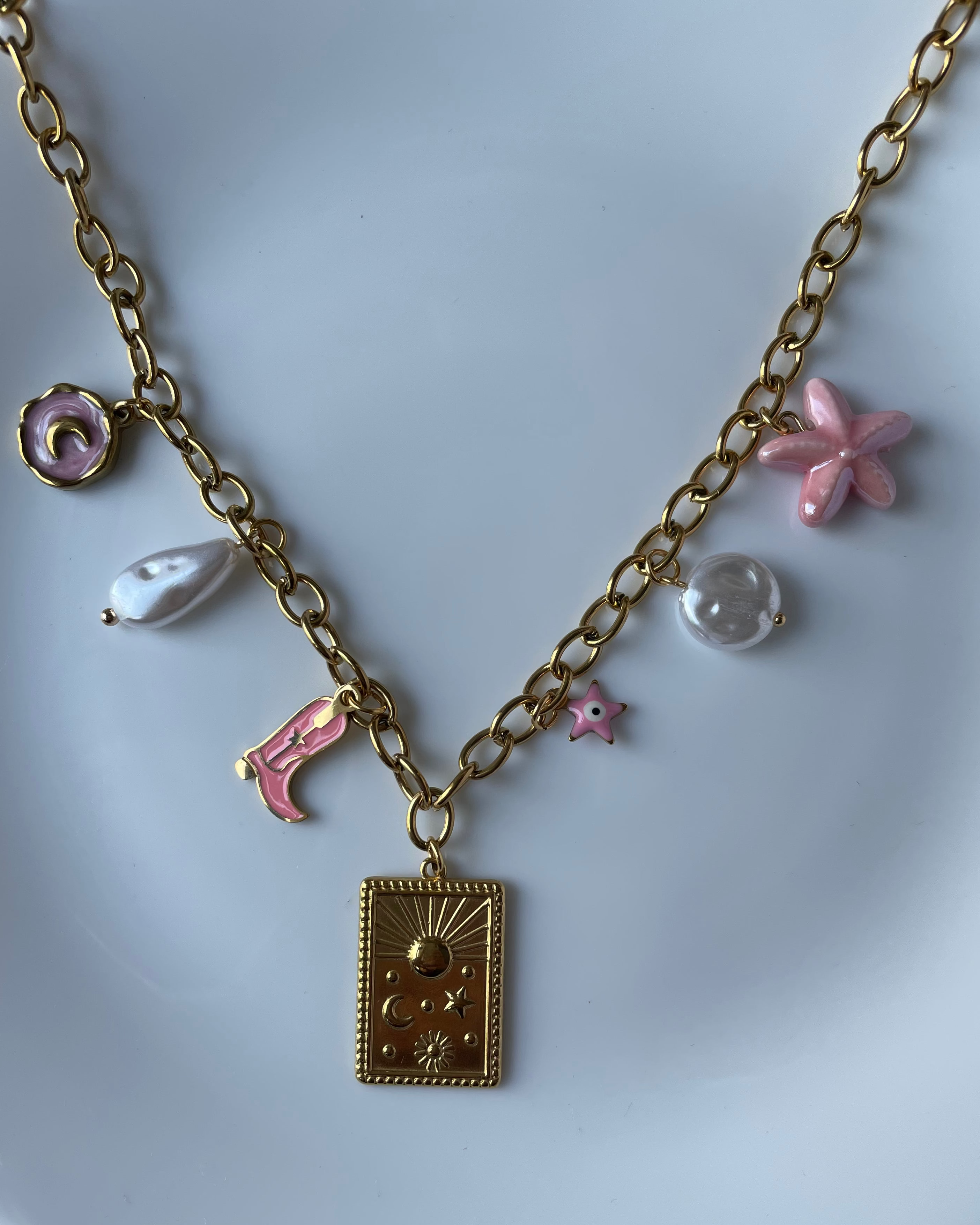 A gold-tone chain necklace with a rectangular pendant and several dangling charms, including pink and white pearl-like beads, displayed on a white plate.