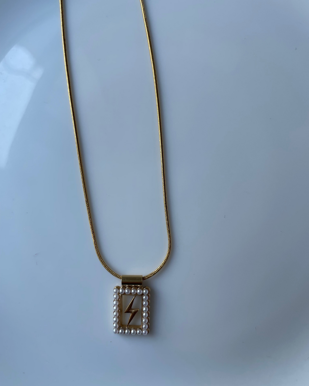 A gold chain necklace with a rectangular pendant featuring a gold lightning bolt in the center, surrounded by a border of small white pearls, displayed on a light blue-white surface.
