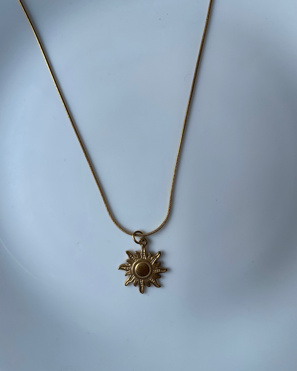 A gold-toned sun pendant necklace on a matching chain, displayed against a light blue background.
