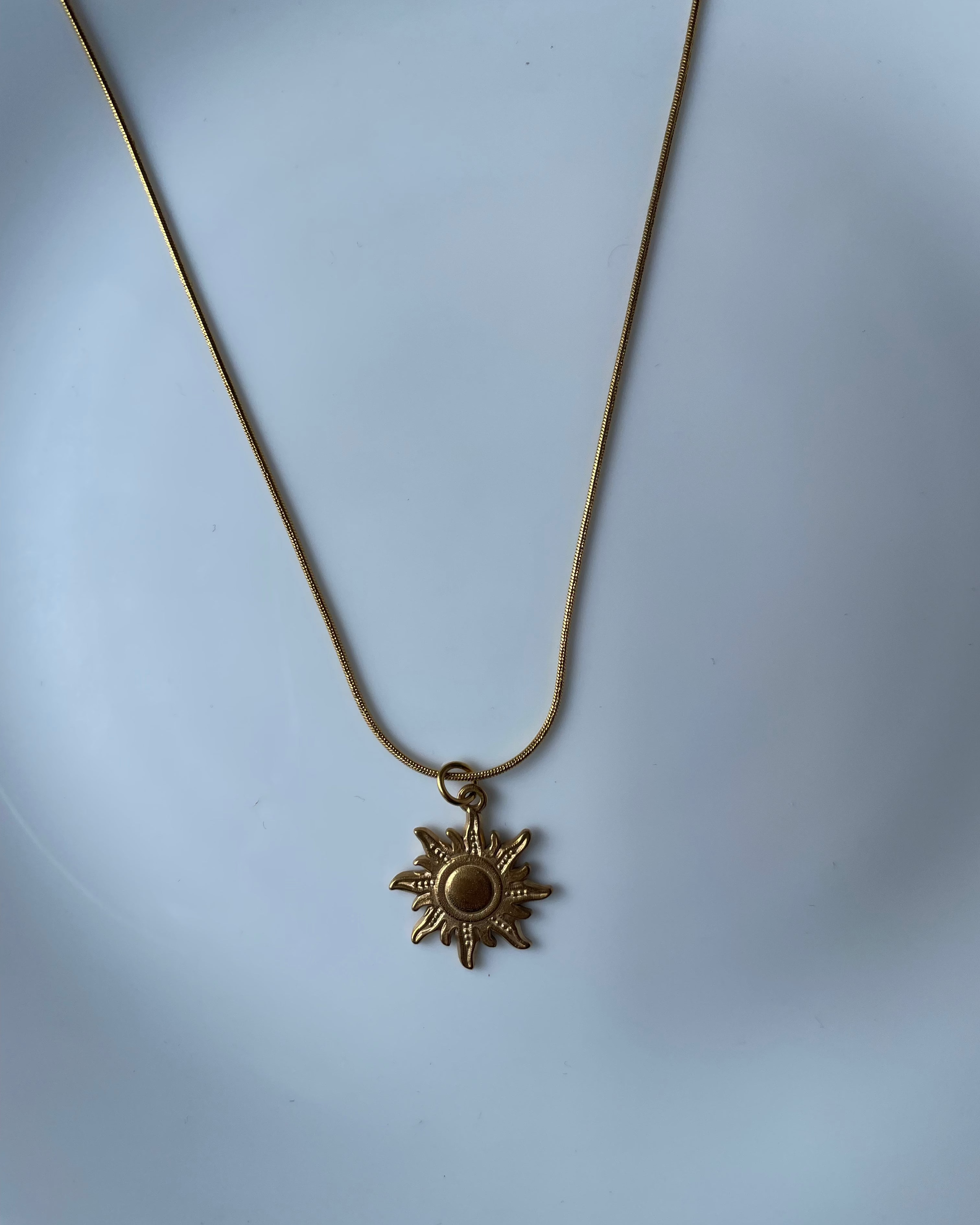 A gold-toned sun pendant necklace on a matching chain, displayed against a light blue background.