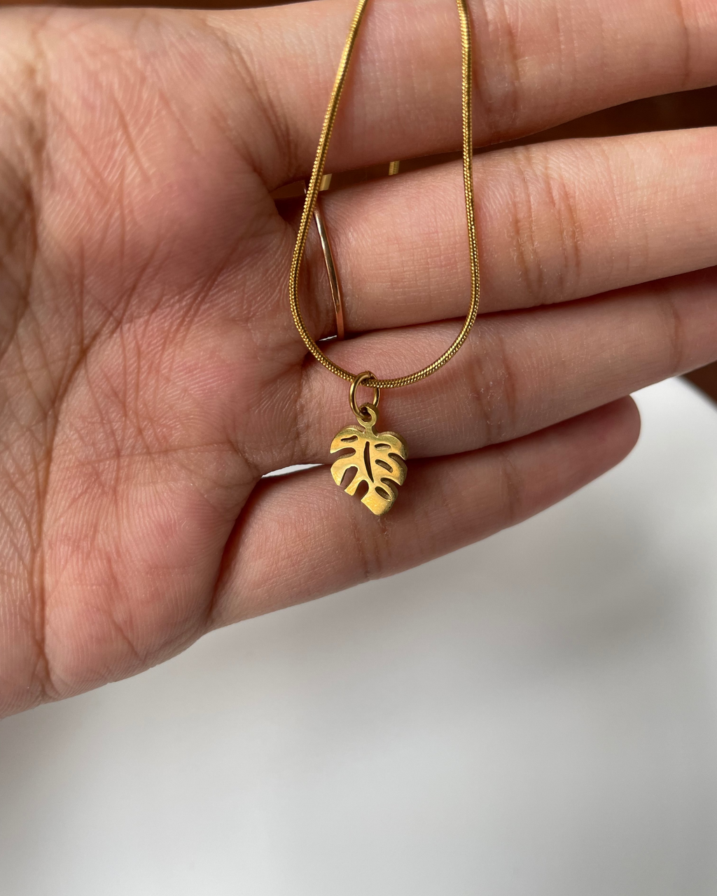 A gold necklace with a small monstera leaf pendant is held in a person's hand against a soft white background.
