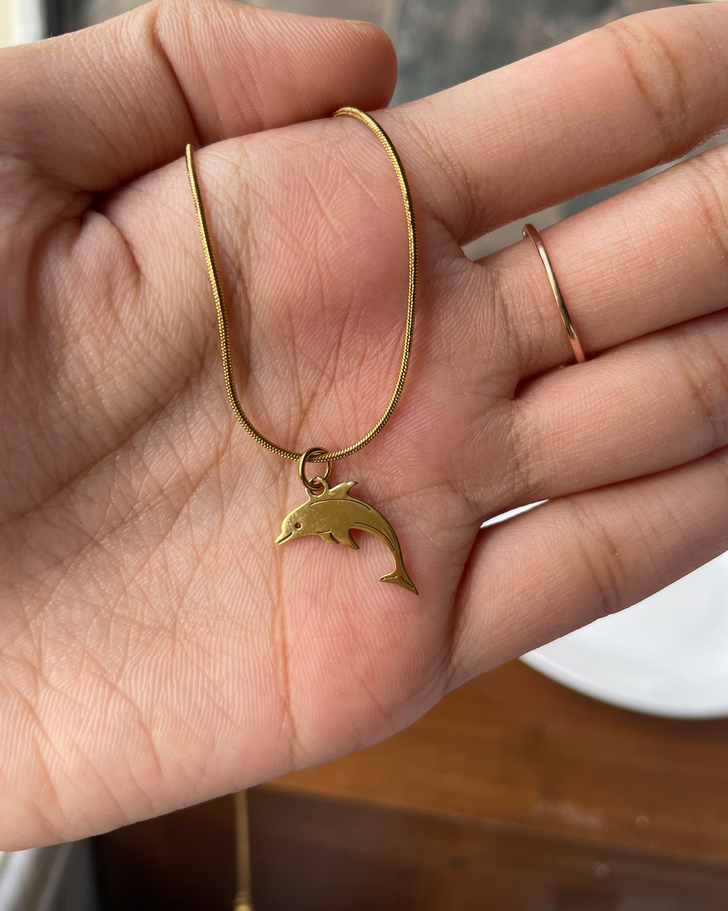A gold-toned snake chain necklace with a small dolphin pendant, held in a person's hand. The person is also wearing a slim rose gold ring on their finger.