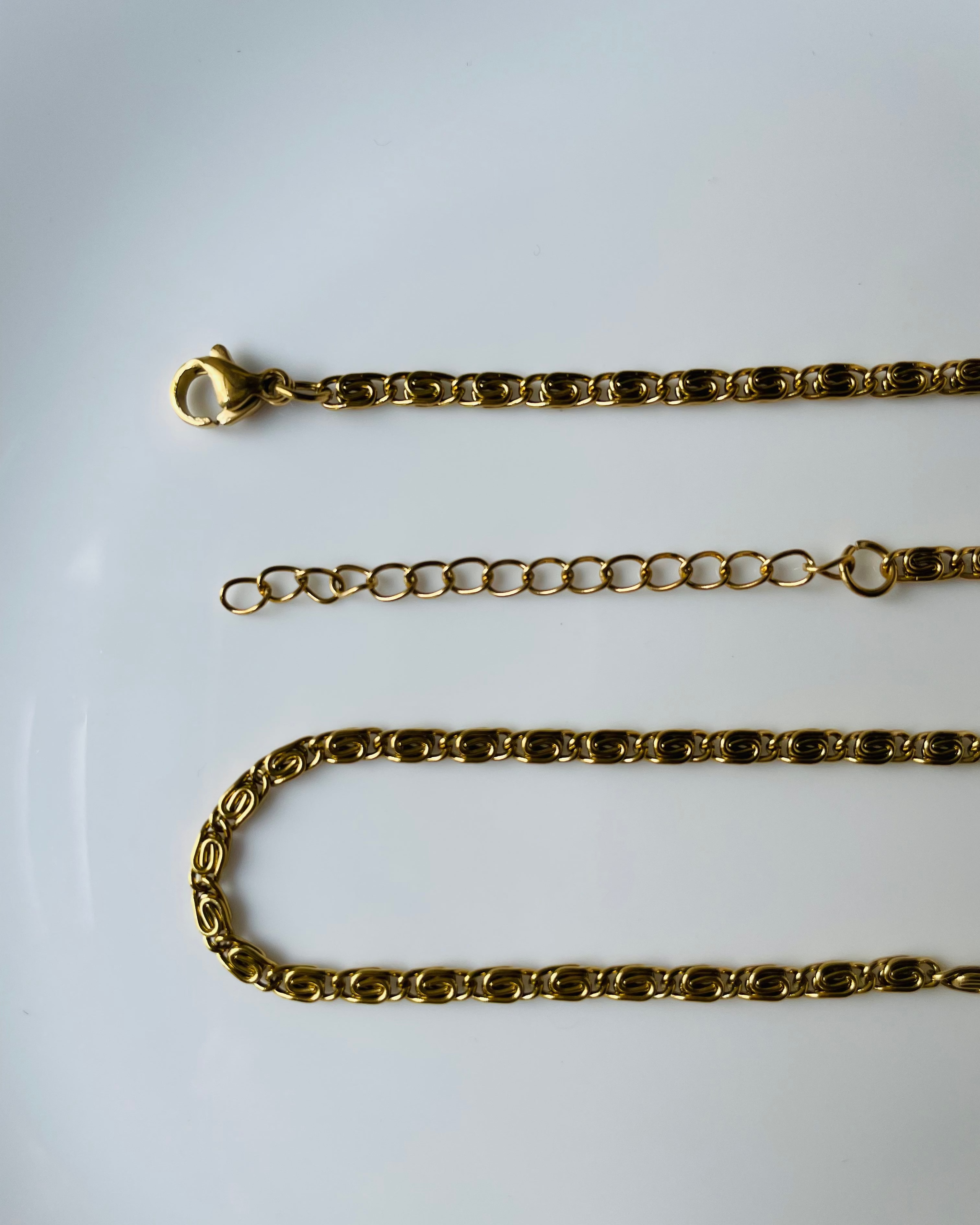 A gold-toned metal chain necklace with a lobster clasp, shown laid out on a plain white surface. The chain includes a short extender section and a longer curb-style link chain.