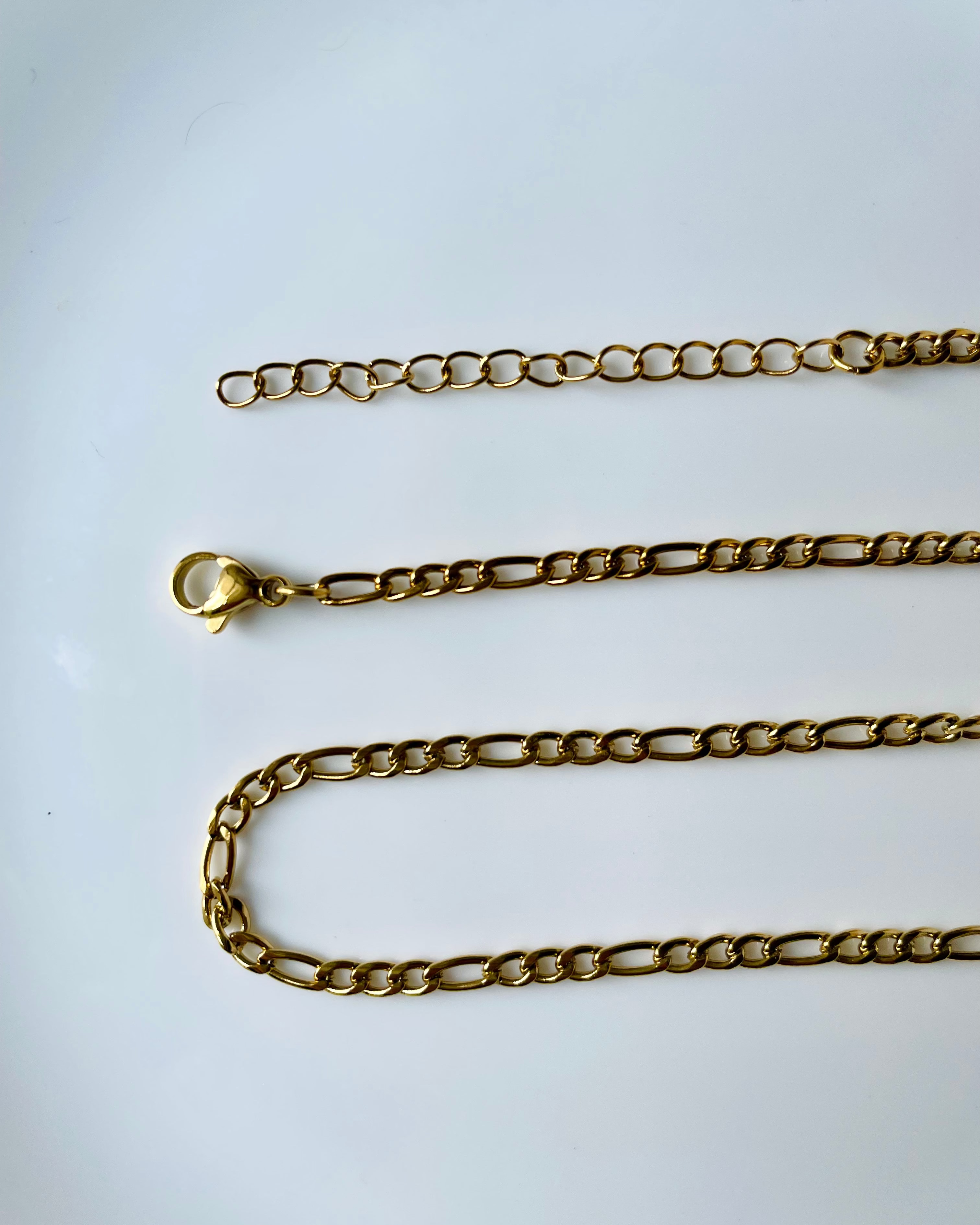 A gold Figaro chain necklace with a lobster clasp, laid out on a plain white surface.