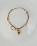 A gold chain bracelet with a small heart charm, displayed on a white plate over a dark wooden surface.