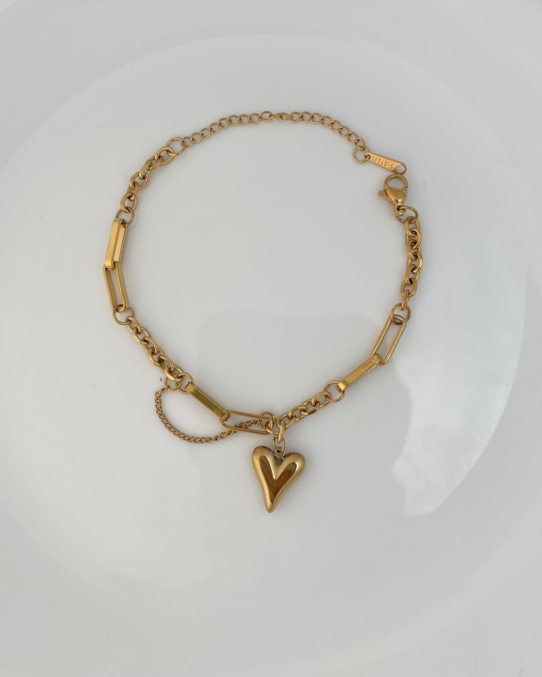 A gold chain bracelet with a small heart charm, displayed on a white plate over a dark wooden surface.