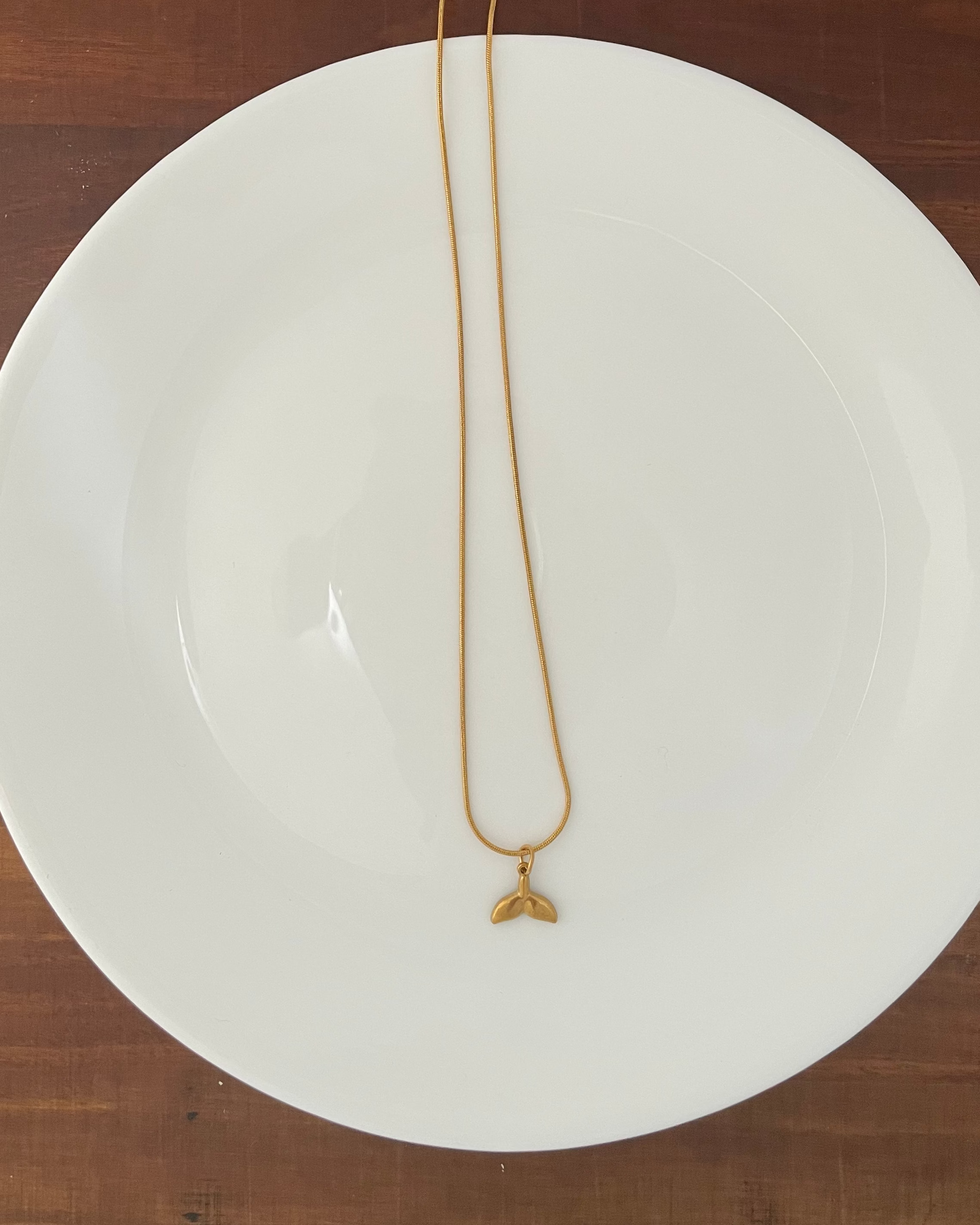 A gold chain necklace with a small gold pendant resting on a white ceramic plate, which is placed on a dark brown wooden table.