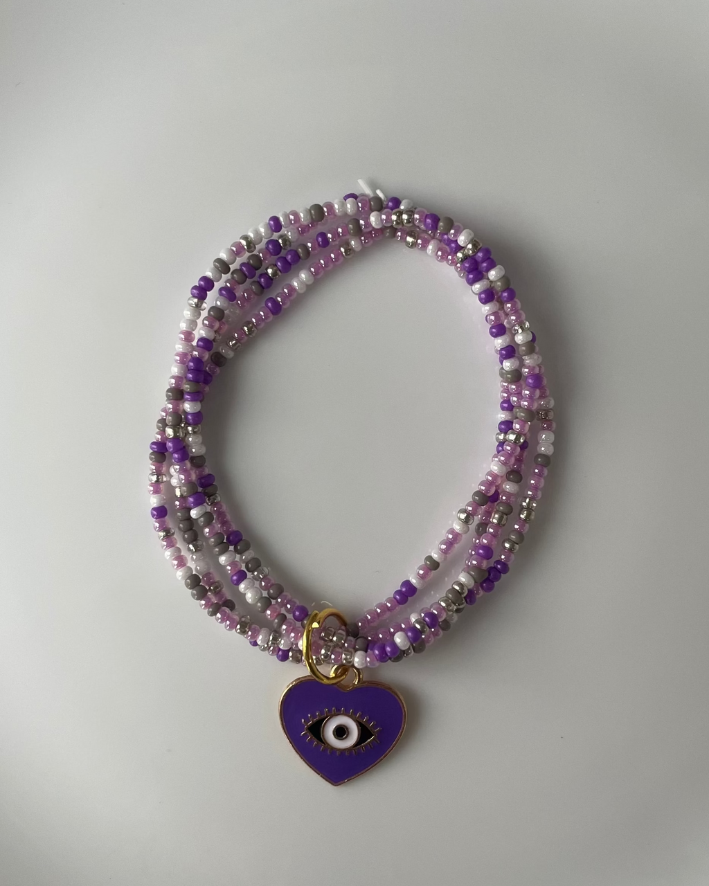 A multi-strand beaded bracelet with purple and silver beads, featuring a small purple heart-shaped evil eye pendant, displayed on a white plate.