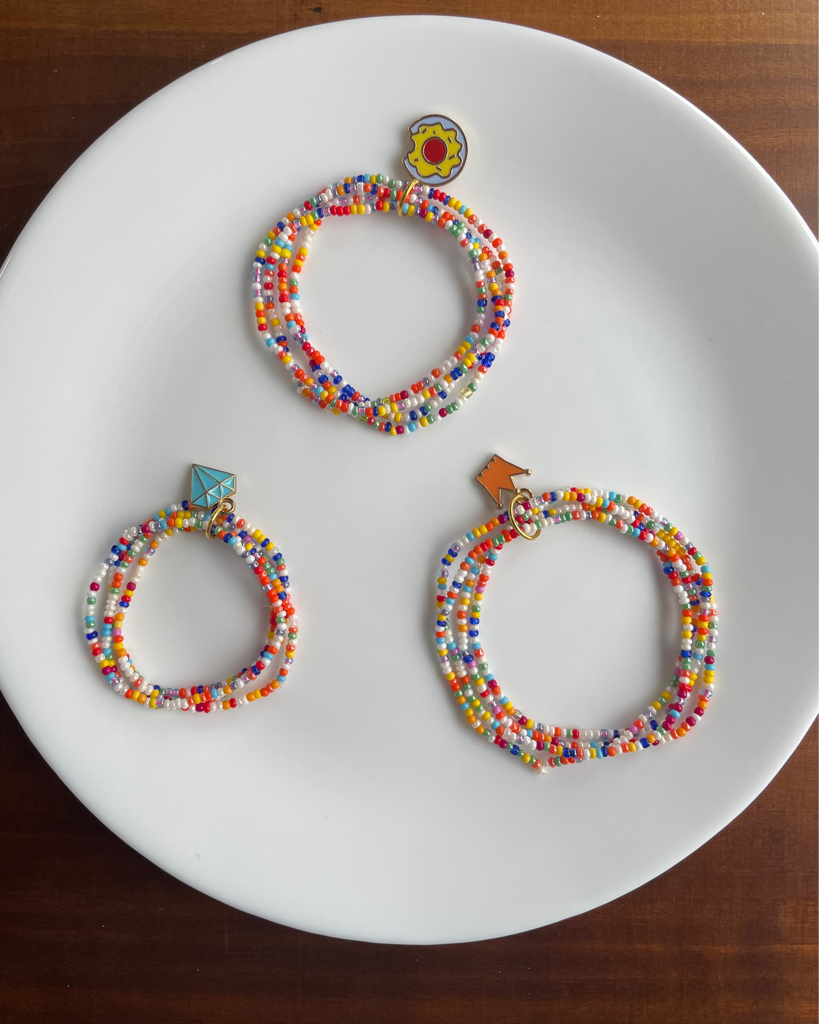 Three colorful beaded hoop earrings with small decorative charms, arranged on a white plate over a dark wooden surface.