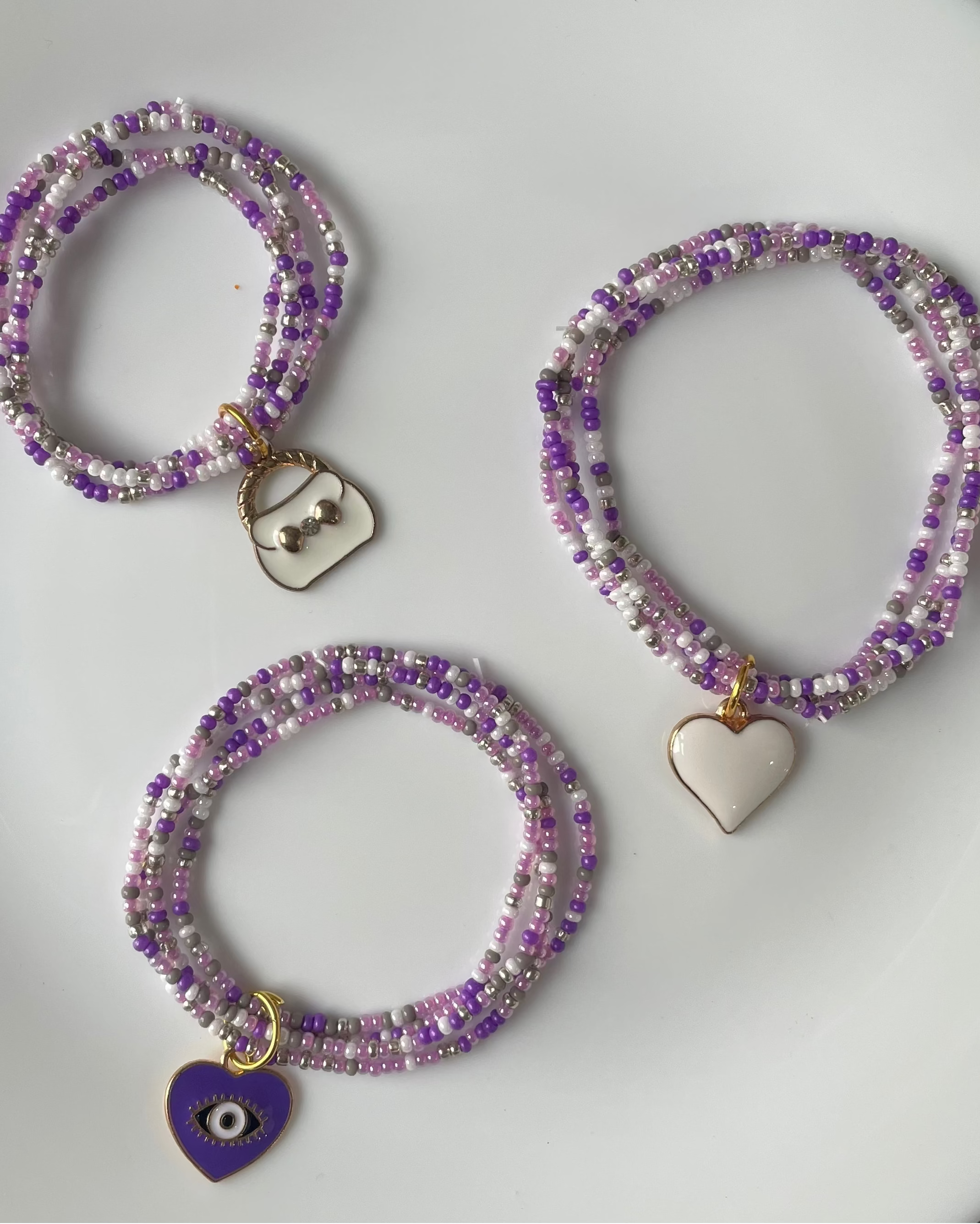 Three beaded bracelets with purple, silver, and white beads are arranged on a white plate. One bracelet has a silver heart charm, one has a white heart charm, and one has a purple evil eye charm.